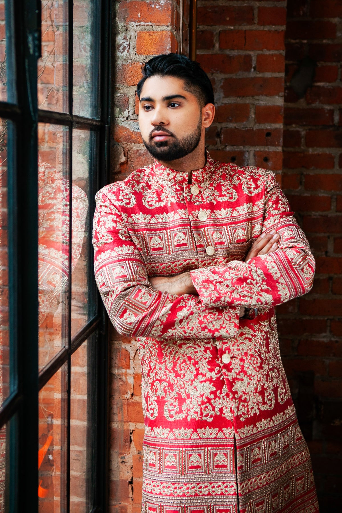 Side view- man wearing a red and gold pattern sherwani leaning on a window with brick walls